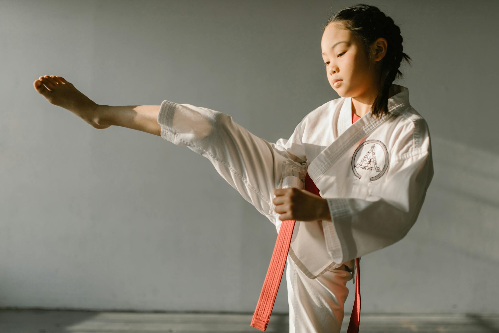 Focused young girl practicing taekwondo kick with red belt indoors.